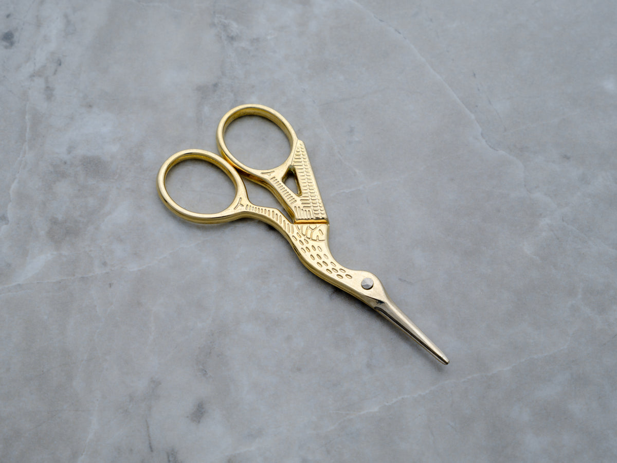 A pair of gold-toned precision trim scissors designed in the shape of a crane. The body of the bird forms the handles, while the long, slender beak serves as the sharp, stainless steel blades, finished in a high-polish gold for a luxury aesthetic.