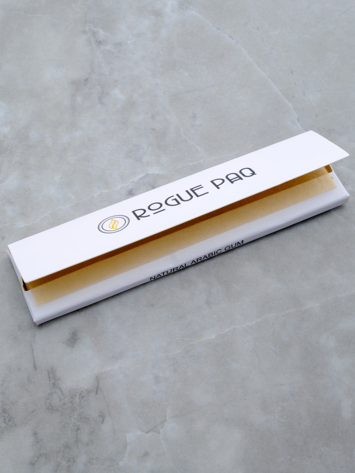 A lead shot of the Rogue Paq Signature King Slim rolling papers. The booklet features a sleek white cover with minimalist black branding. A single, unbleached brown hemp paper is pulled forward, showcasing its natural, additive-free texture and king slim dimensions