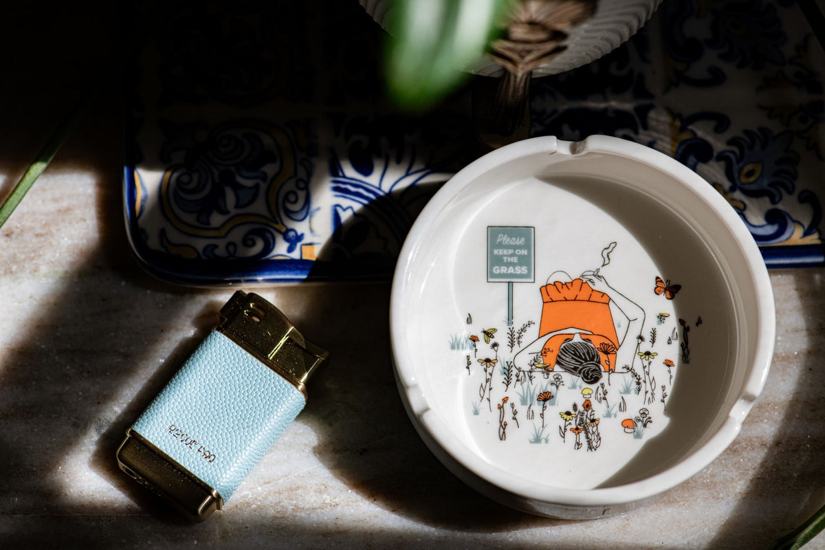 White porcelain ashtray with intricate detail featuring a sign that says Please Keep ON The Grass. The ashtray is styled with the Rogue Paq refillable lighter in sky blue.