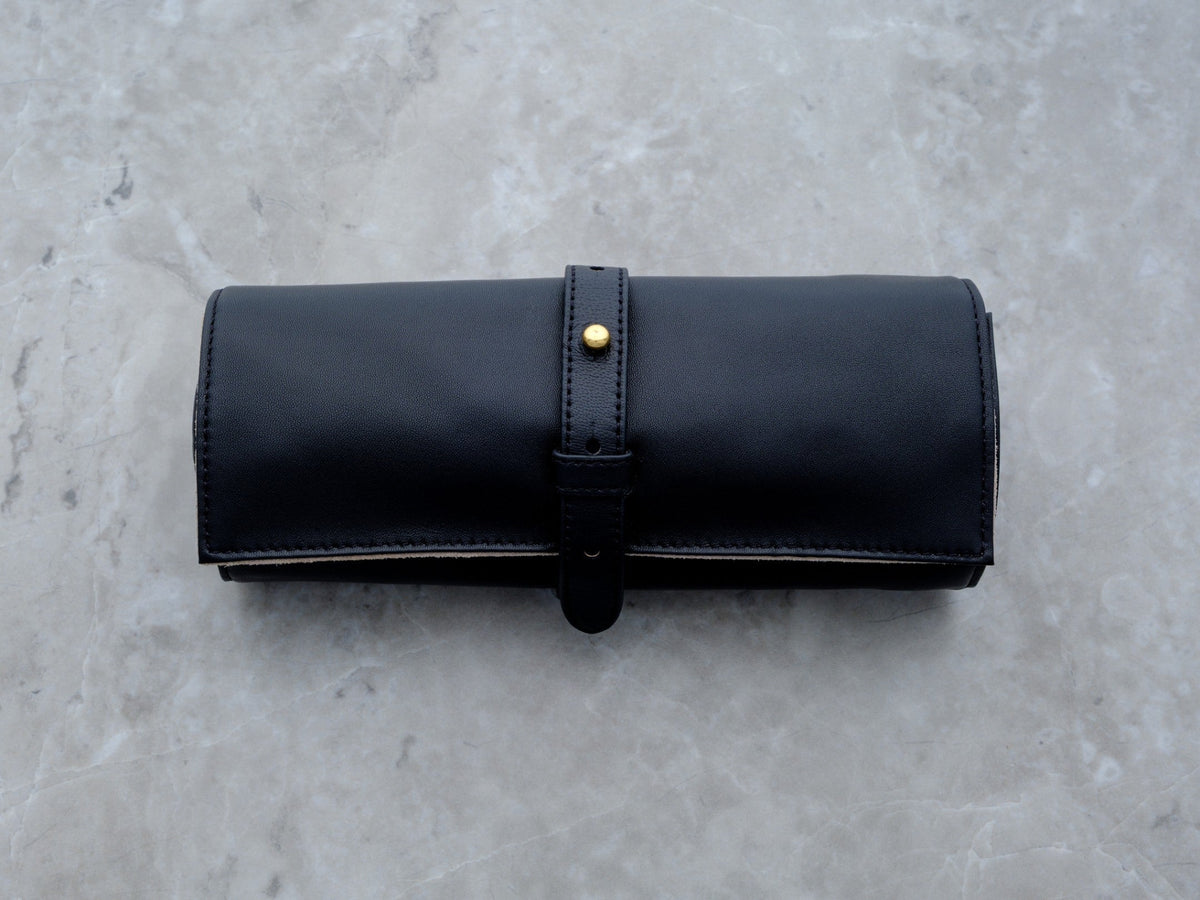 A clean, horizontal studio shot of the black lambskin Ritual Case. The uniform grain of the leather and the minimalist gold-toned hardware are showcased against a gray marble background, highlighting its high-fashion aesthetic.