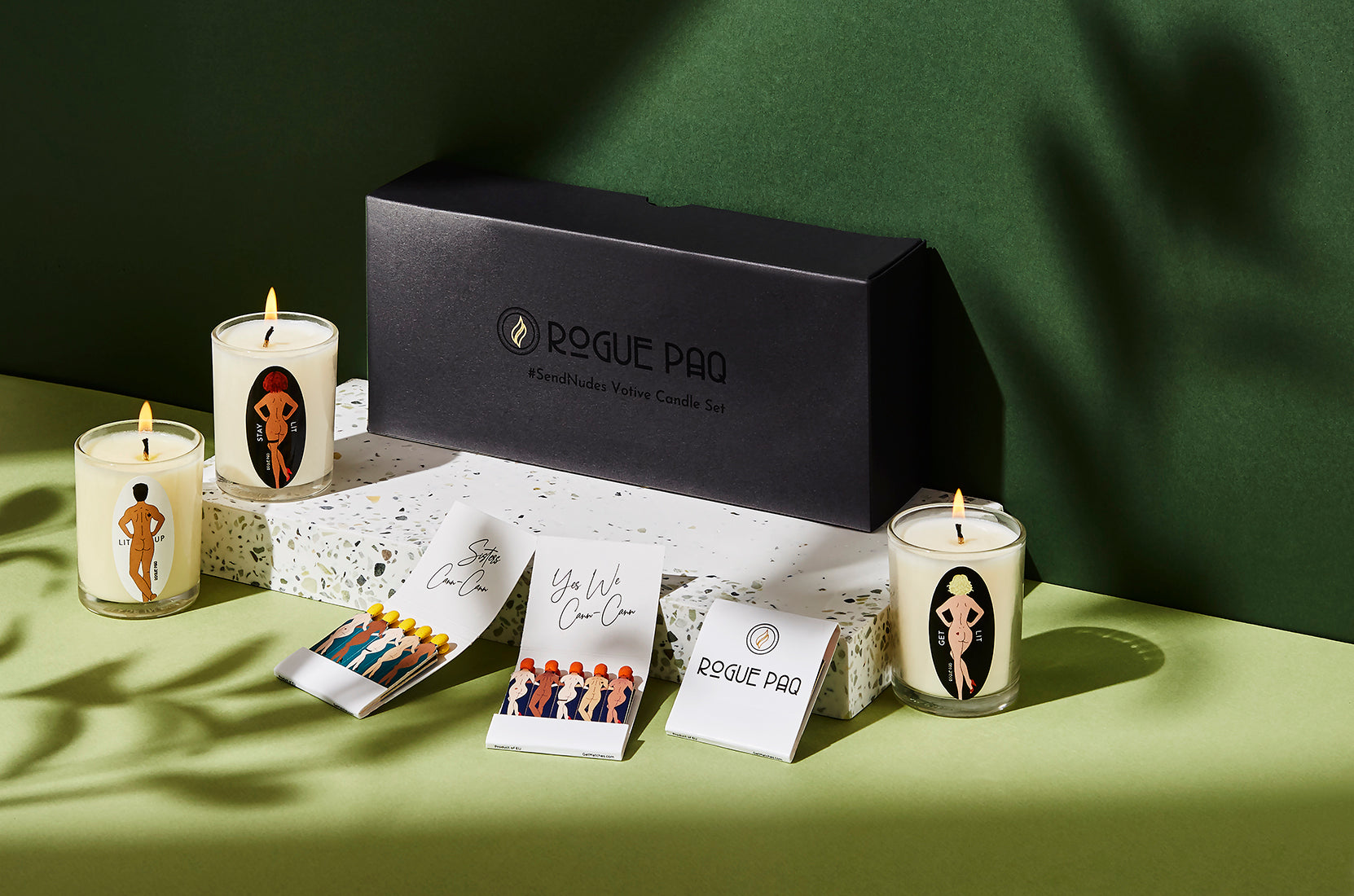 A detailed shot of three soy wax aromatherapy candles plue Rogue Paq designer matchbooks from the SendNudes collection. The 'Sisters Cann-Cann' and Yes We Cann Cann illustrations feature diverse, artistic nude figures with varying skin tones and hair colors in a high-kick dance pose, representing a celebration of body positivity and inclusivity. The shot includes the sleek minimalist matte black gift box.