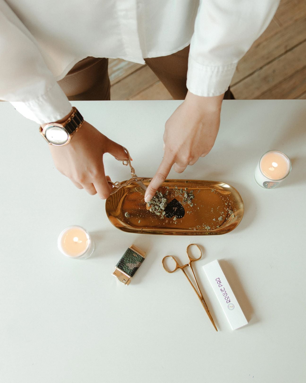 The Joie de Weed tray in use, showing its perfect scale for rolling a joint. The smooth stainless steel surface and slightly curved edges are designed for ease of use, ensuring no flower is wasted during the ritual. It is pictured with a refillable lighter, two rogue paq massage candles, a book of all natural hemp rolling papers, and rogue paq hemostat joint clips.
