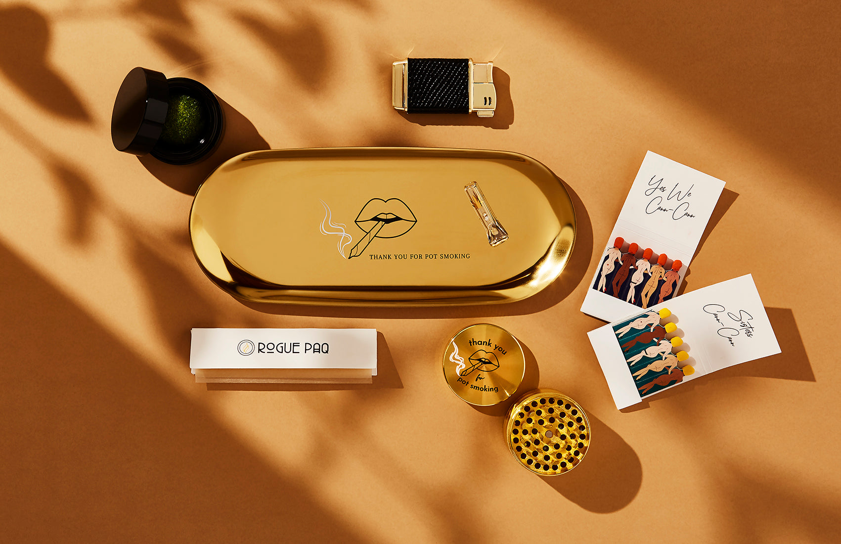 Gold-tone joint rolling tray with black 'Thank You for Pot Smoking' retro graphic; displayed with a matching 4-piece gold herb grinder for a cohesive aesthetic. Also includes Rogue Paq SendNudes matches, and premium rolling papers along with a refillable lighter; all essential accessories are included in the Thank You Ritual Gift Set