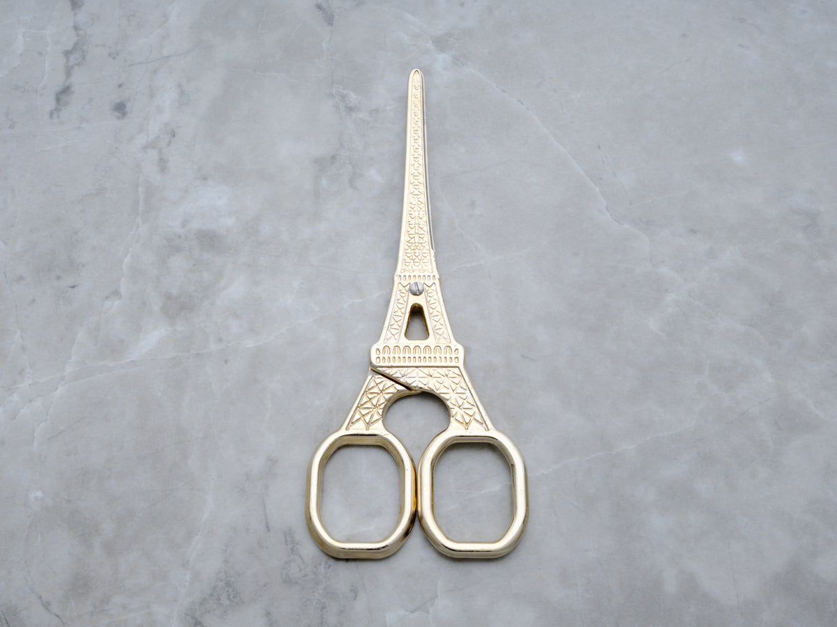 A flat lay shot of the scissor handles, highlighting the intricate latticework and architectural details of the Eiffel Tower design. The high-polish gold finish catches the light, emphasizing the boutique quality of this Rogue Paq accessory.