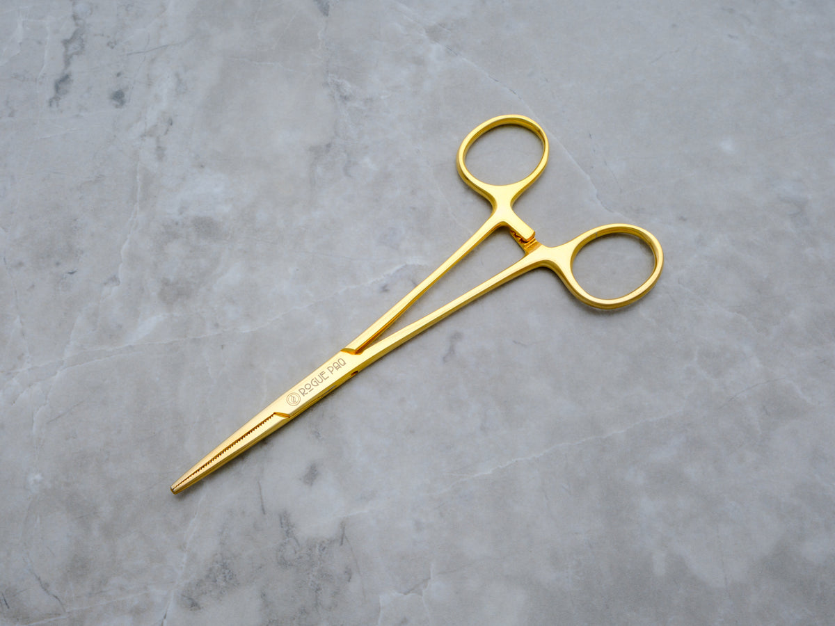 A pair of Rogue Paq gold-toned hemostat jay-clips. The surgical-grade stainless steel tools feature a high-shine gold finish and a locking mechanism, repurposed as a sophisticated accessory for packing or holding hand-rolled joints.