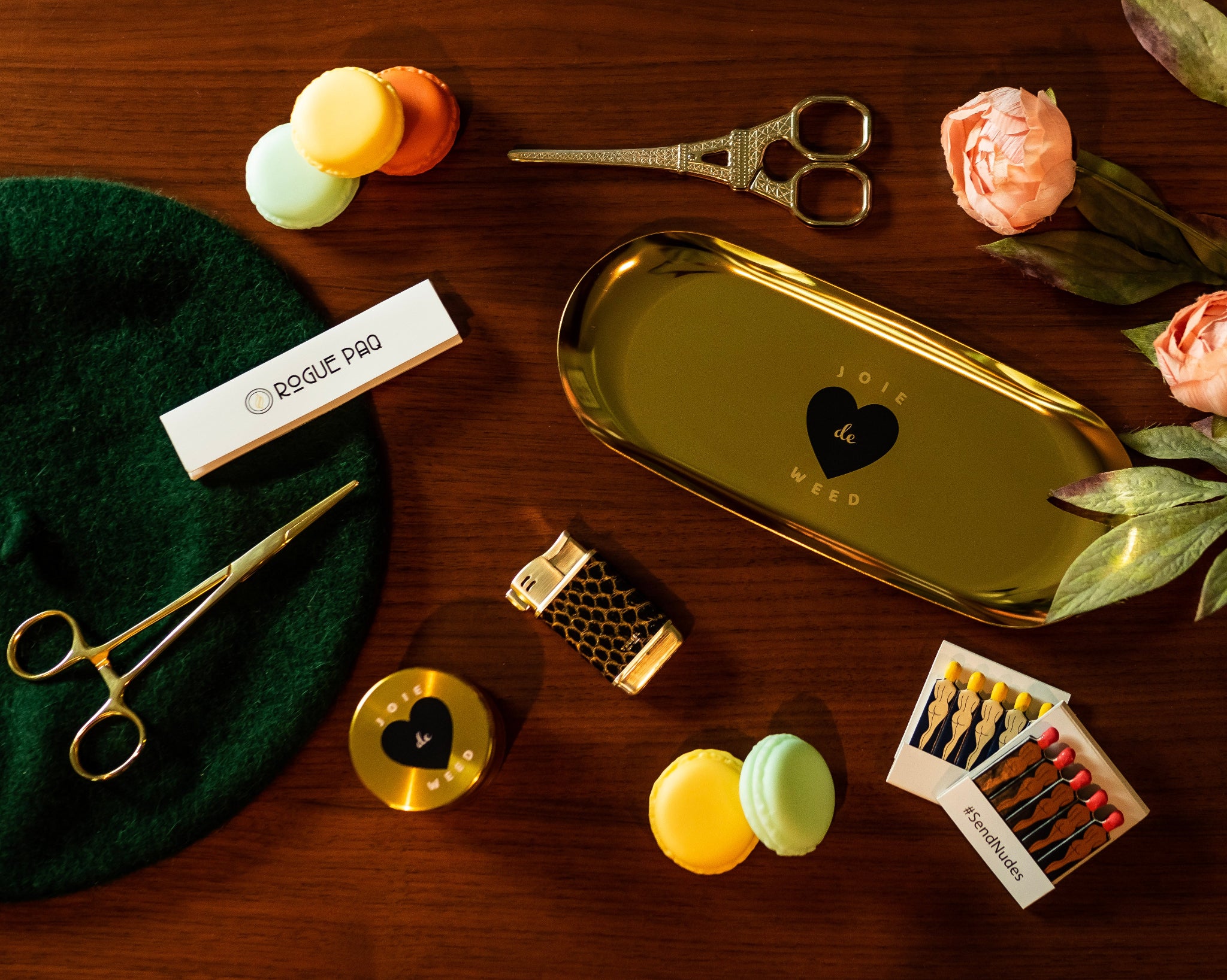 A luxury cannabis gift set featuring the gold 50mm 'Joie de Weed' 4-piece grinder, a booklet of Rogue Paq unbleached hemp rolling papers, Eiffel Tower Scissors, a refillable lighter, two books of SendNudes matches, and a pair of gold-toned hemostat jay clips. The set is presented in a minimalist, high-contrast arrangement on a wooden surface that highlights the polished gold hardware. Macarons and a beret are in the shot to emphasize the Parisian vibes.