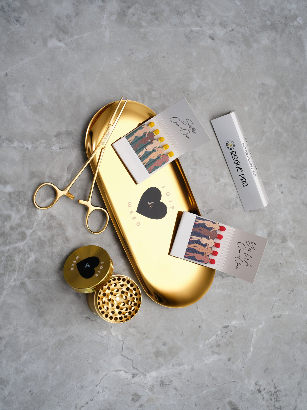 A luxury cannabis gift set featuring a booklet of Rogue Paq unbleached hemp rolling papers, Eiffel Tower Scissors, a refillable lighter, two books of SendNudes matches, and a pair of gold-toned hemostat jay clips. The set is presented in a minimalist, high-contrast arrangement that highlights the polished gold hardware.