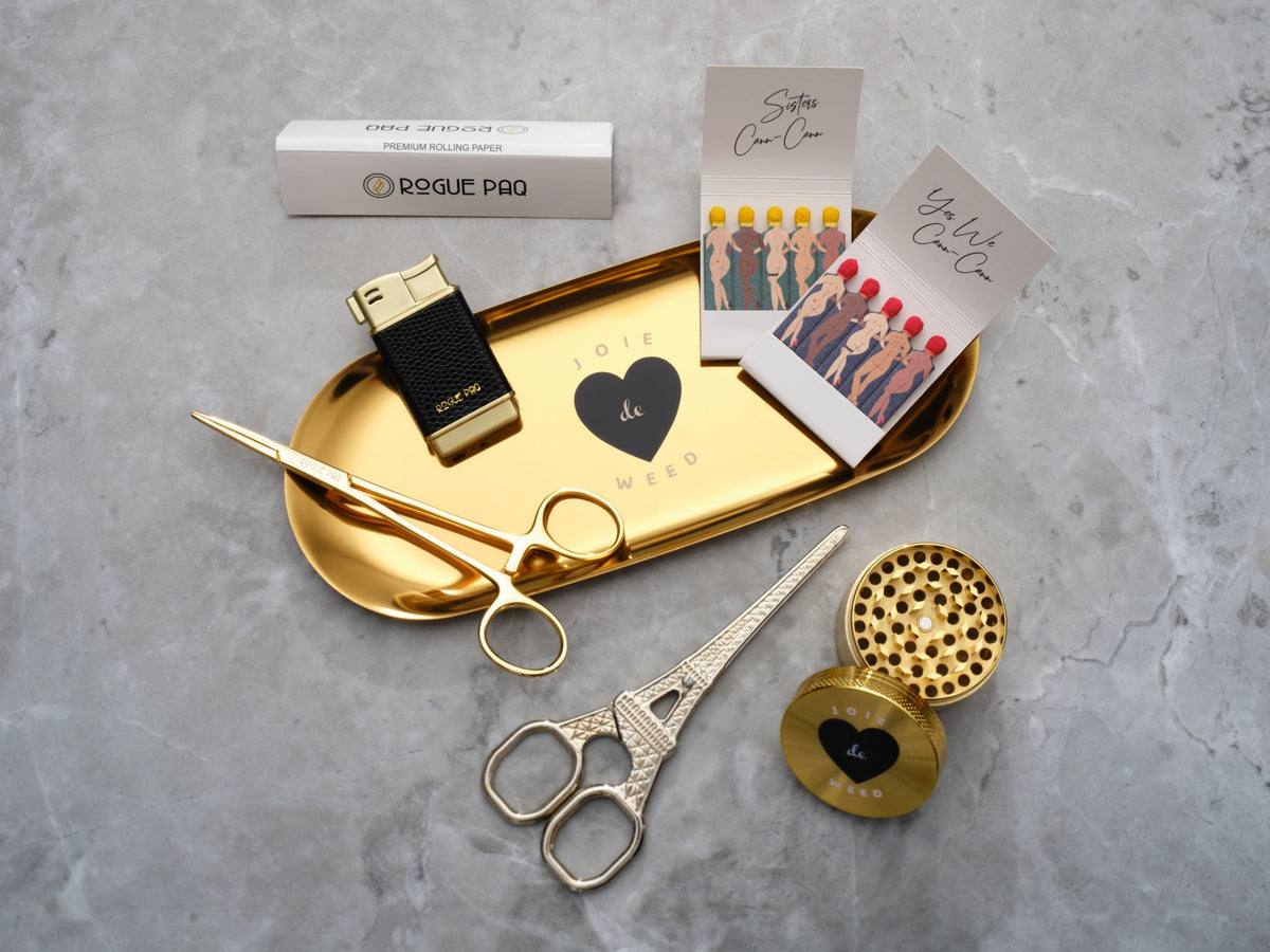A luxury cannabis gift set featuring a booklet of Rogue Paq unbleached hemp rolling papers, Eiffel Tower Scissors, a refillable lighter, two books of SendNudes matches, and a pair of gold-toned hemostat jay clips. The set is presented in a minimalist, high-contrast arrangement that highlights the polished gold hardware.