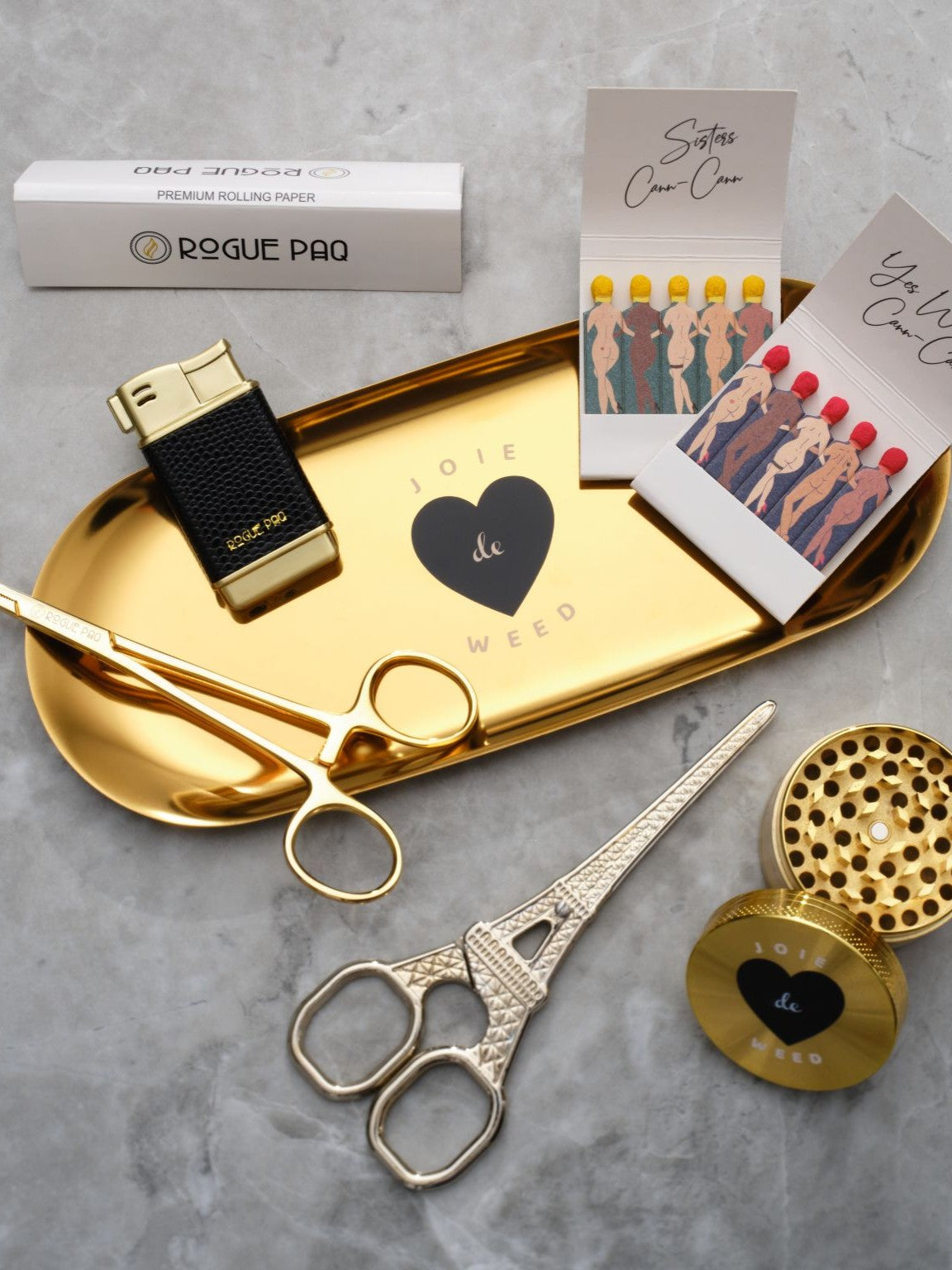A luxury cannabis gift set featuring a booklet of Rogue Paq unbleached hemp rolling papers, Eiffel Tower Scissors, a refillable lighter, two books of SendNudes matches, and a pair of gold-toned hemostat jay clips. The set is presented in a minimalist, high-contrast arrangement that highlights the polished gold hardware.