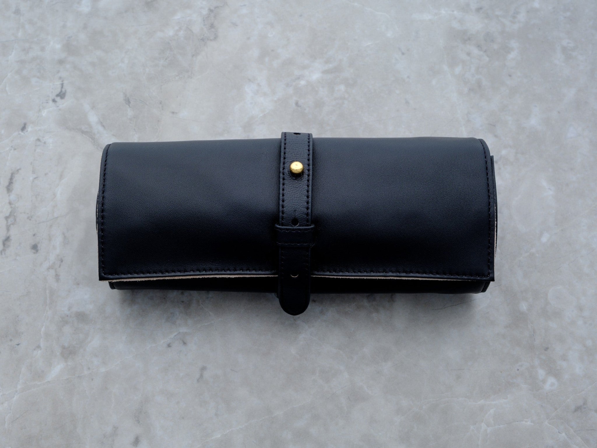 A clean, horizontal studio shot of the black lambskin Ritual Case. The uniform grain of the leather and the minimalist gold-toned hardware are showcased against a gray marble background, highlighting its high-fashion aesthetic.