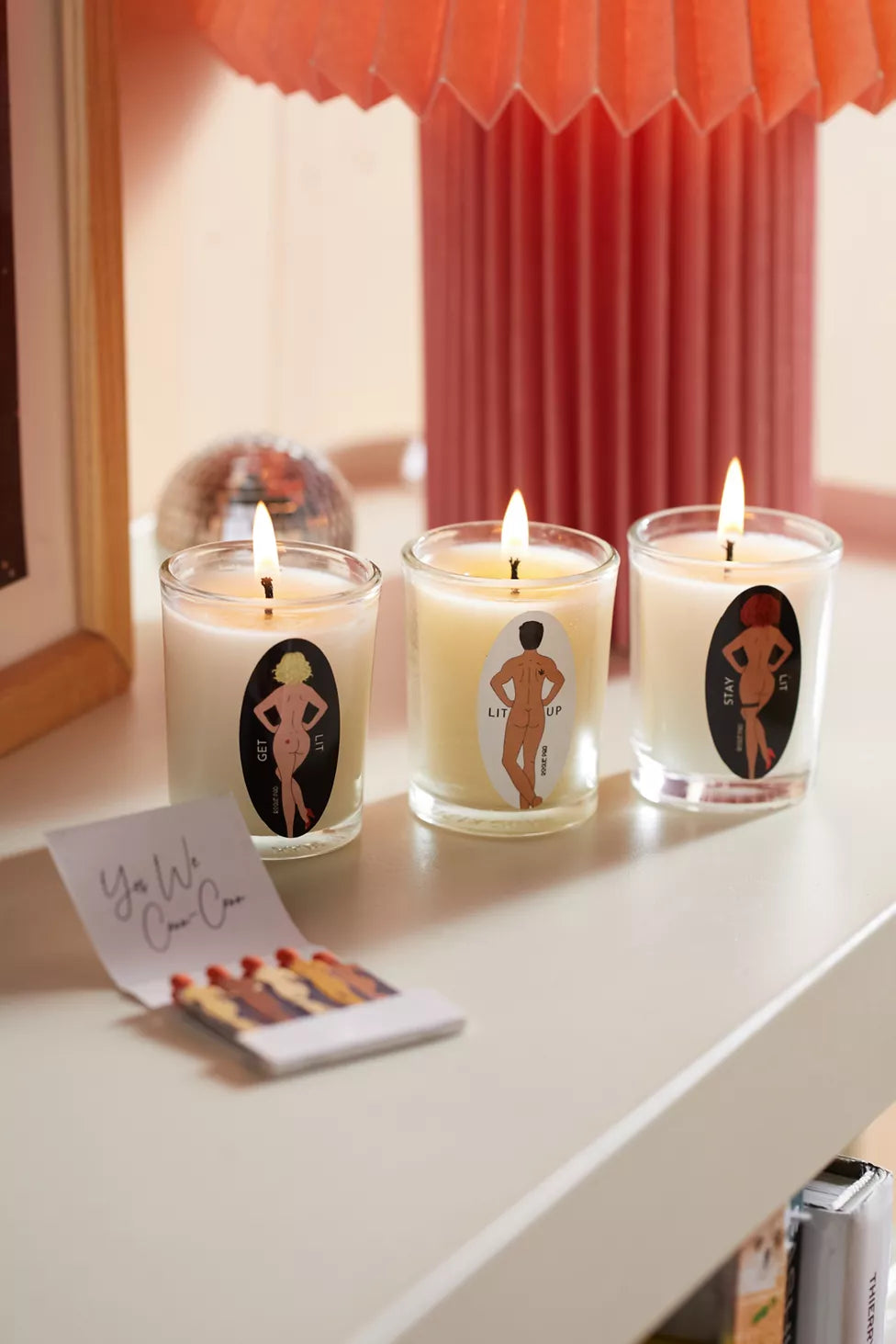 Three lit candles with artistic labels featuring the Signature SendNudes dancing figures on a white surface, with a peach lamp in the background and an open Yes We Cann Cann matchbook in front of the candles.
