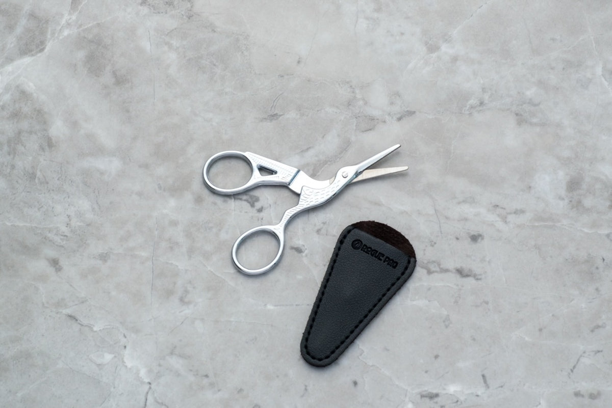 Rogue Paq intricate crane scissors in silver displayed with its vegan blade cover on a marble background.