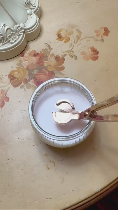 The gold wick trimmer being used to maintain a clear glass candle. The image demonstrates the precision and ease of the tool, emphasizing the Rogue Paq philosophy of 'Raising Your Ritual' through proper maintenance and high-design hardware.