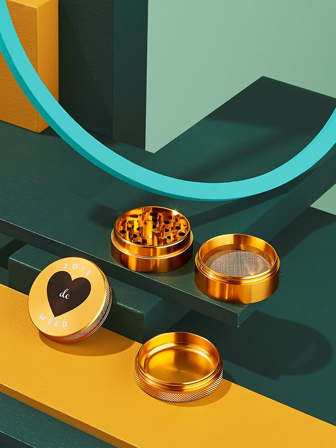 An exploded view of the Joie de Weed gold grinder, displaying the magnetic lid with diamond-shaped teeth, the grinding chamber, the fine stainless steel sifting screen, and the bottom kief collection chamber. The interior is polished for smooth operation and easy maintenance.