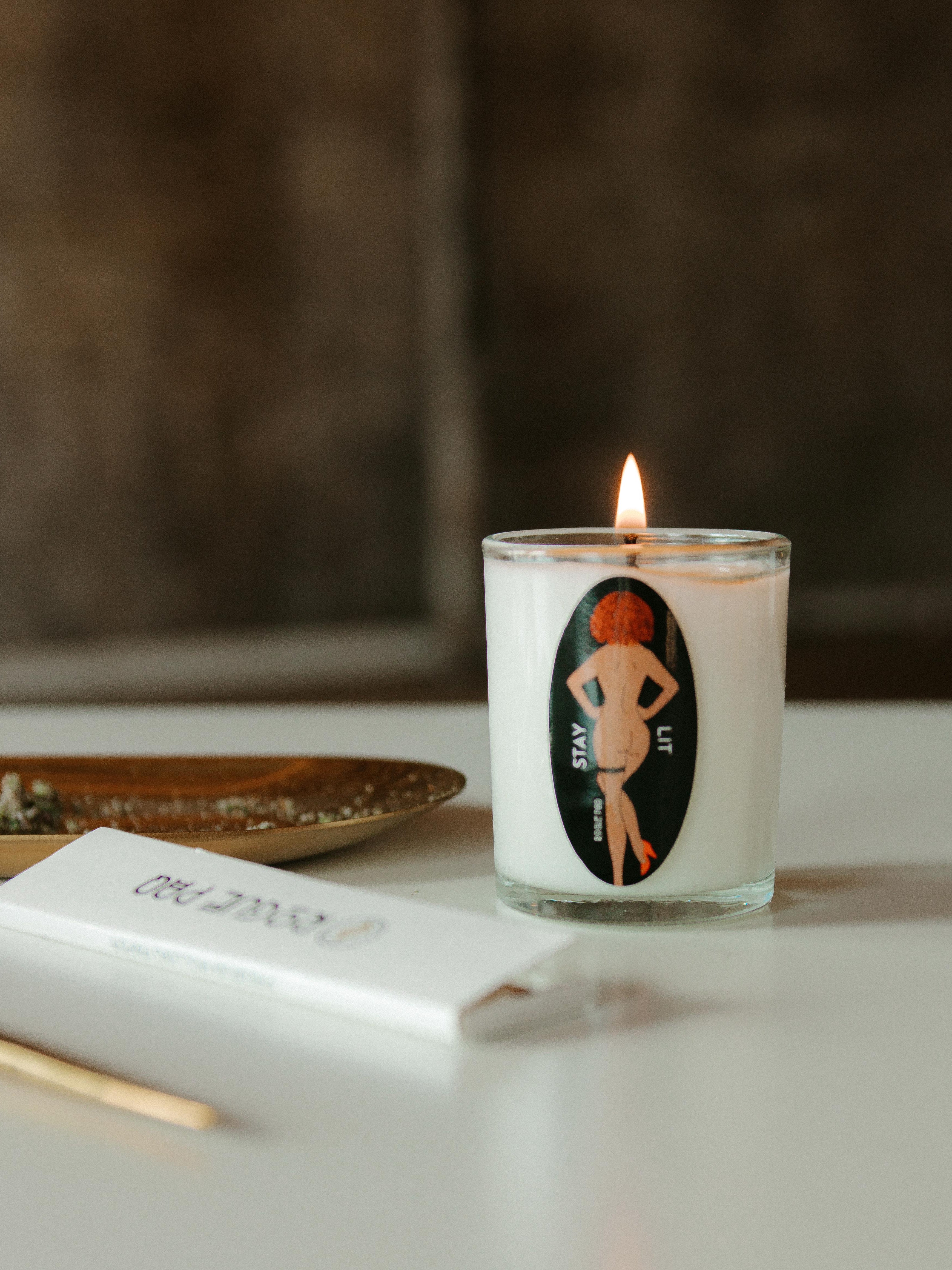 A side-by-side shot of the STAY LIT organic massage candle in a clear glass votive and a book of Rogue Paq all natural hemp rolling papers. The candle features a minimalist black label featuring their signature SendNudes nude dancer with 'STAY LIT' in a sophisticated black serif font. The label displays the 'SendNudes' illustration of one artistic figure with brown skin and red hair in a high-kick dance pose.